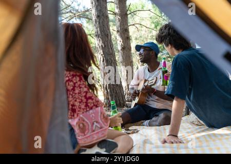 Eine Gruppe junger, vielfältiger Freunde, die in einem Wald campen, wobei einer Gitarre spielt, während andere zuhören. Das Bild fängt einen Moment der Freizeit und des Zusammenlebens ein Stockfoto