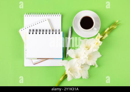 Künstlerische Komposition mit geöffnetem leerem Notizblock und Stift, Tasse schwarzen Kaffee, Blumen eines weißen Gladiolen auf grünem Hintergrund. Festliches Desktop-Konzept für das Büro. Kaffeetasse am Morgen. Stockfoto