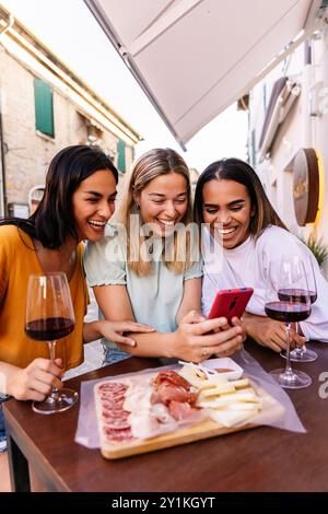 Drei junge Frauen, die ihr Handy benutzen, während sie einen Aperitif in einer Bar genießen Stockfoto