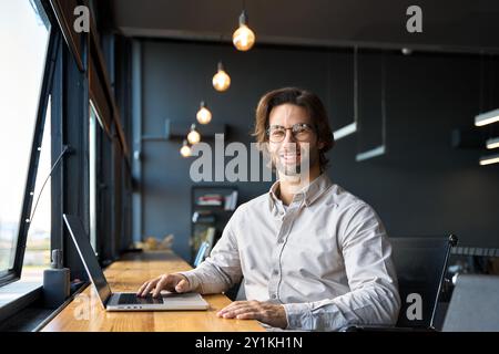 Glücklicher junger Geschäftsmann, der im Büro die Kamera ansieht, die am Laptop arbeitet. Stockfoto