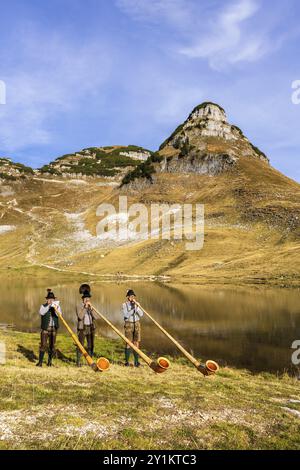 Drei Männer spielen das Alphorn am Augstsee am Mount Loser. Das österreichische Alphorntrio Klangholz. Der Atterkogel im Hintergrund. Herbst, gutes Wetter Stockfoto