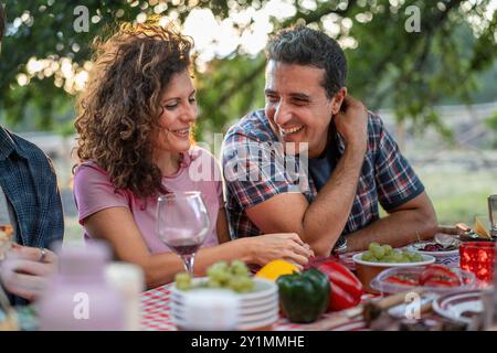 Fröhliches Paar mittleren Alters, das sich bei einer Dinner-Party im Freien einen intimen Moment teilt. Romantische Picknickszene mit Wein und frischen Produkten. Liebe, Beziehungen und Stockfoto