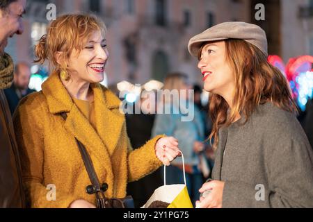 Zwei stilvolle Frauen, die den Abend in der Stadt genießen und lebhafte Gespräche führen. Freunde, die zusammen einkaufen, Tasche halten, in trendigen Wintermänteln gekleidet. Stockfoto