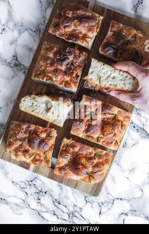 Eine Hand, die ein Stück frisch gebackenes Focaccia-Brot von einem Holzbrett pflückt. Das Brot ist goldfarben, mit Rosmarinzweigen und Salzflocken belegt und hat eine Stockfoto