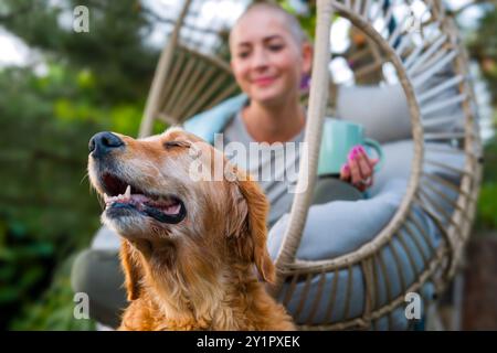 Junge Krebspatientin entspannt lächelnd im Hängesessel auf der Terrasse mit ihrem emotionalen Unterstützungshund an ihrer Seite. Das psychische Wohlbein Stockfoto