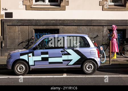 Frau in rosafarbenen und gelben Kleidern, die neben einem auffällig bemalten kleinen Auto steht, Köln, Deutschland. Frau in rosa und gelbe Kleidung steht neben einem auf Stockfoto