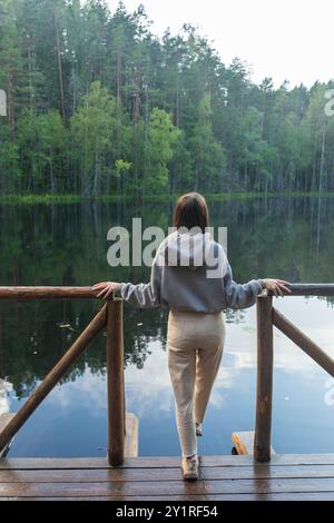 Junge Frau, die auf einem hölzernen Dock steht, mit Blick auf einen ruhigen See, umgeben von einem dichten Wald. Das ruhige Wasser reflektiert das üppige Grün und schafft ein Stockfoto