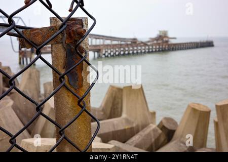 Colwyn Bay, North Wales, Großbritannien, 09-08-2024. Rostiger Zaun mit einem Pier am Meer im Hintergrund, der einen Kontrast zwischen industriellem und natürlichem Element schafft Stockfoto