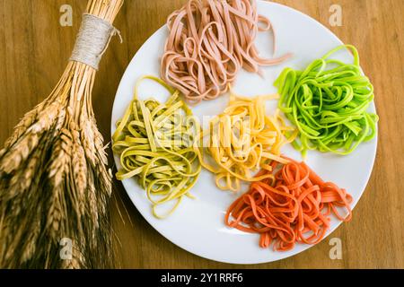 Italienische Pasta in verschiedenen Farben auf einem weißen Teller auf einem Holztisch mit einem Haufen Weizen. Gekochte mehrfarbige Tagliatelli. Hochwertige Fotos Stockfoto