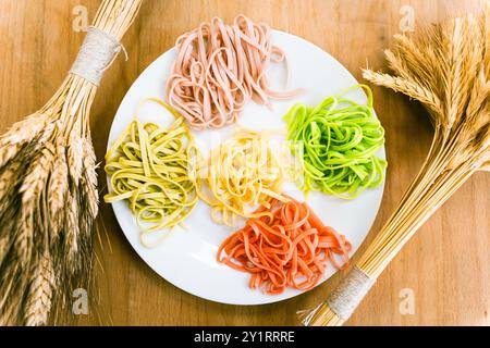 Italienische Pasta in verschiedenen Farben auf einem weißen Teller auf einem Holztisch mit einem Haufen Weizen. Gekochte mehrfarbige Tagliatelli. Hochwertige Fotos Stockfoto