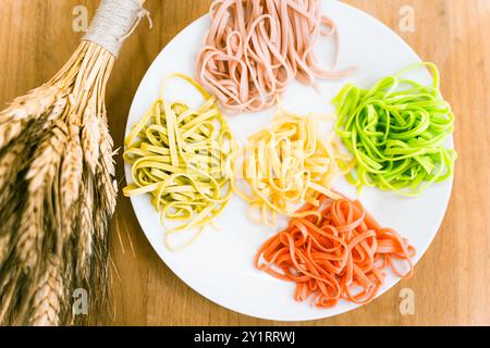 Italienische Pasta in verschiedenen Farben auf einem weißen Teller auf einem Holztisch mit einem Haufen Weizen. Gekochte mehrfarbige Tagliatelli. Hochwertige Fotos Stockfoto