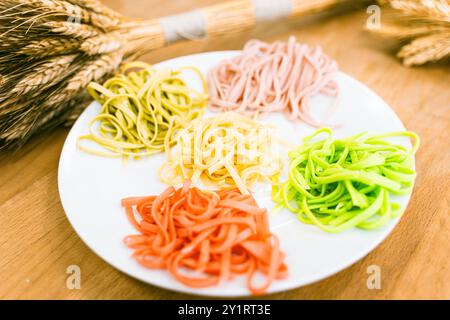 Italienische Pasta in verschiedenen Farben auf einem weißen Teller auf einem Holztisch mit einem Haufen Weizen. Gekochte mehrfarbige Tagliatelli. Hochwertige Fotos Stockfoto