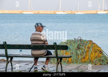 Ein alter Mann sitzt allein am Hafen in zakynthos oder der Stadt zante in griechenland, ein pensionierter Mann sitzt auf einer Bank neben bunten Fischernetzen in zante. Stockfoto