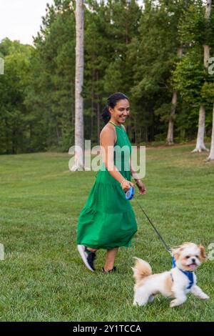 Eine Frau in einem grünen Kleid geht mit ihrem Hund in einem Park. Der Hund ist ein kleiner weißer Pudel. Die Frau lächelt und scheint ihre Zeit mit ihm zu genießen Stockfoto