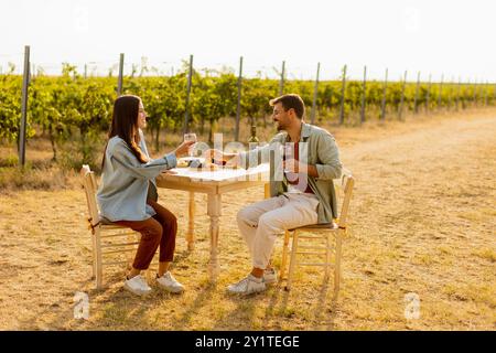 Ein paar entspannen sich an einem rustikalen Tisch in einem sonnigen Weinberg, genießen Wein und Gourmet-Snacks. Sie teilen Lachen und Freude, umgeben von üppigen Weinstöcken und dem Stockfoto