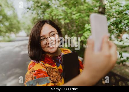 Fröhliche junge Dame mit Brille, die ein Selfie in einem üppig grünen Garten macht. Fröhliche Frau mit Smartwatch und Laptop, die an einem sonnigen Tag ein Selfie macht Stockfoto