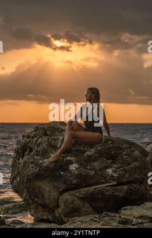 Eine Frau im Badeanzug sitzt auf einem Felsen am Meer und blickt auf den Sonnenuntergang. Goldenes Licht der Sonne durchdringt die Wolken. Stockfoto