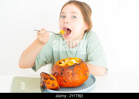 Süßes Mädchen isst Kürbissuppe, serviert in gebackenem Kürbis. Glückliches Mädchen in einem grünen T-Shirt mit Herbstschale für Halloween auf weißem Hintergrund Stockfoto