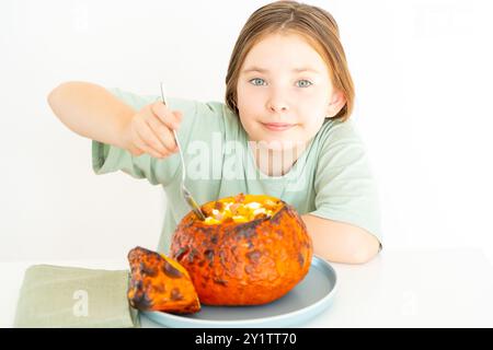 Süßes Mädchen isst Kürbissuppe, serviert in gebackenem Kürbis. Glückliches Mädchen in einem grünen T-Shirt mit Herbstschale für Halloween auf weißem Hintergrund Stockfoto