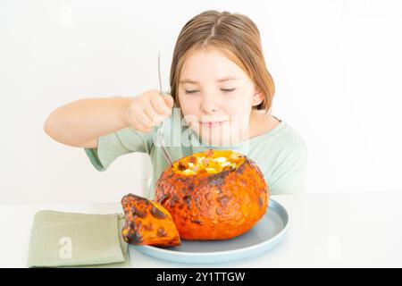 Kleines Mädchen im Teenageralter isst Kürbissuppe, serviert in gebackenem Kürbis. Mädchen in grünem T-Shirt und Herbstschale auf weißem Hintergrund. Hochwertige Pho Stockfoto