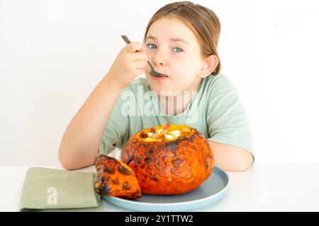 Süßes Mädchen isst Kürbissuppe, serviert in gebackenem Kürbis. Glückliches Mädchen in einem grünen T-Shirt mit Herbstschale für Halloween auf weißem Hintergrund Stockfoto