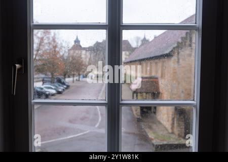 Ein regnerischer Blick durch ein Fenster, mit Tropfen auf dem Glas, zeigt eine entfernte verschwommene Szene einer alten Straße mit geparkten Autos und Schloss langenburg Stockfoto