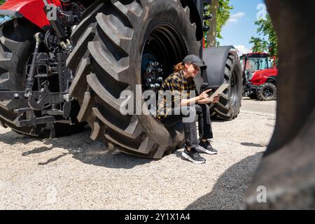 Eine Landwirtin sitzt auf dem großen Rad eines landwirtschaftlichen Traktors und nutzt ihr digitales Tablet. Stockfoto