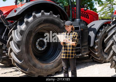 Eine Farmerin steht neben einem großen Rad eines Traktors und benutzt ihr digitales Tablet. Stockfoto