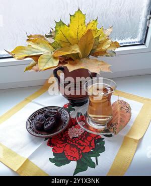 Herbstzeit und Herbst kreative Motive zu Hause. Stillleben im Herbst. Ahornblätter in Keramikkrug (Ton) und ein Glas Tee mit Pflaumen Stockfoto