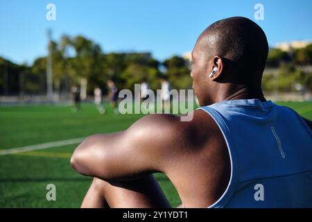 Junge Sportmannsfußballspieler tragen kabellose Ohrhörer, blaues athletisches Tanktop, das auf einem grasbewachsenen Sportfußballfeld sitzt und nach dem Spiel oder Workou ausruht Stockfoto