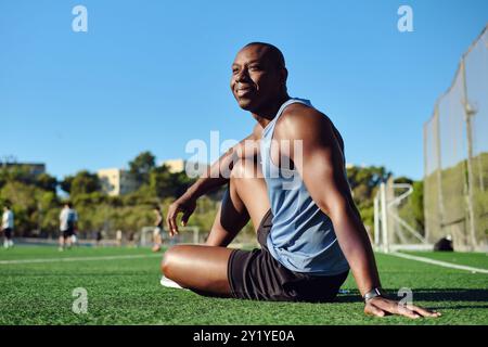 Sportlicher Mann Fußballspieler tragen blaues, sportliches Tanktop und schwarze Shorts, sitzen auf einem grasbewachsenen Fußballfeld, ruhen sich nach dem Spiel oder Workout aus, sehen aus Stockfoto
