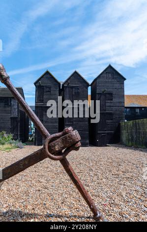 Drei schwarze Fischernetzschuppen in Rock-a-Nore, Hastings an einem sonnigen Tag mit blauem Himmel und weißen Wolken, mit einem Anker im Vordergrund Stockfoto