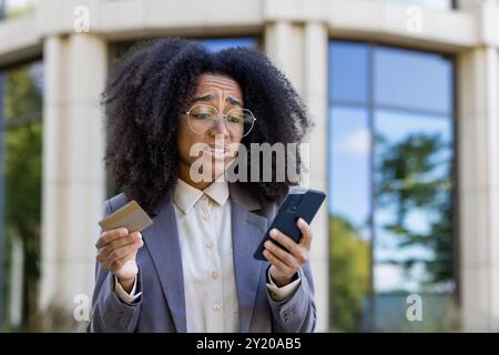 Afroamerikanische Geschäftsfrau, die ihre Besorgnis ausdrückt und Kreditkarte und Smartphone vor einem modernen Bürogebäude hält. Steht für Online-Banking, Finanzmanagement, digitale Transaktionen. Stockfoto