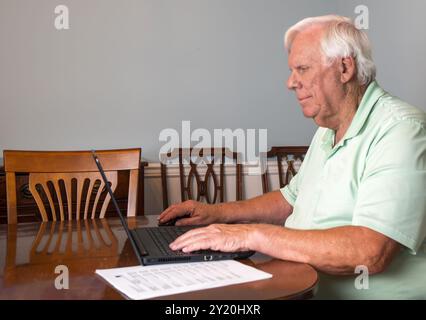 Ein älterer Mann mit weißen Haaren und grünem Hemd arbeitet an seinem Laptop in einem provisorischen Büro in seinem Esszimmer. Stockfoto