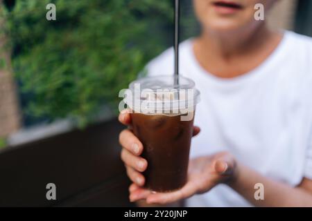 Beschnittener Schuss einer unerkennbaren Frau in weißem T-Shirt, die eine Plastiktasse zum Mitnehmen mit kaltem Kaffeetrink im Freien hält. Stockfoto