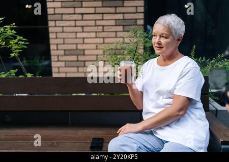 Porträt einer älteren Frau, die Eiskaffee im Café trinkt, zufrieden und entspannt in der Stadt aussieht. Europäisches grauhaariges älteres Weibchen, das auf der Bank sitzt Stockfoto