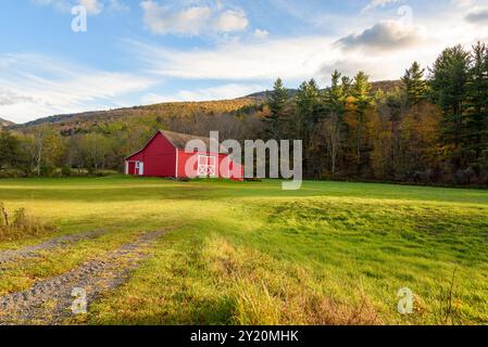 Traditionelle rote amerikanische Holzscheune auf einem grasbewachsenen Feld am Fuße eines bewaldeten Berges bei Sonnenuntergang im Herbst Stockfoto