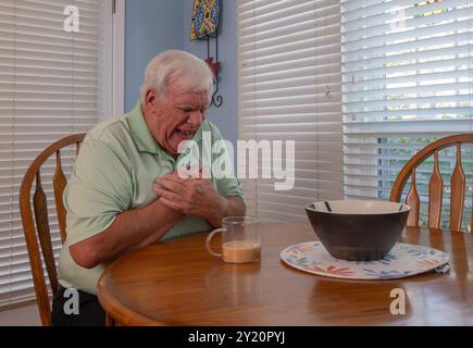 Ein älterer Mann mit weißem Haar, der ein grünes Hemd trägt, genießt seinen Kaffee und schaut aus dem Fenster, wenn er Brustschmerzen hat. Stockfoto