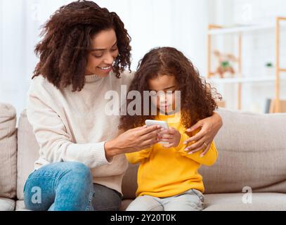 Afrikanische Mutter Unterrichtet Tochter, Um Smartphone Auf Sofa Zu Sitzen Stockfoto