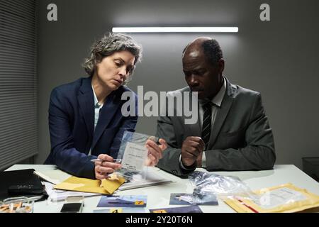 Zwei Detectives untersuchen Beweise und diskutieren entscheidende Aspekte der Untersuchung in hell beleuchtetem Büro, umgeben von gelben Mappen, Umschlägen und Beweisbeuteln Stockfoto