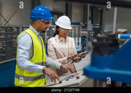 Die indische Betriebsleiterin trainiert einen afroamerikanischen männlichen Mitarbeiter in einem blauen Helm und einer gelben Weste, um Maschinen zu bedienen. Stockfoto