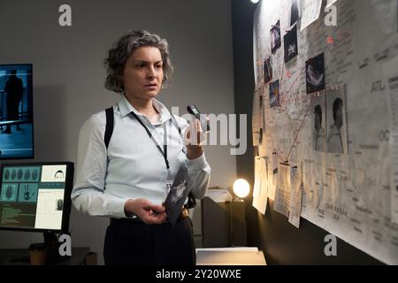 Frau, die Fotos von Kriminalszenen überprüft und Hinweise auf der Evidenztafel analysiert, während sie die Taschenlampe im Büro hält Stockfoto