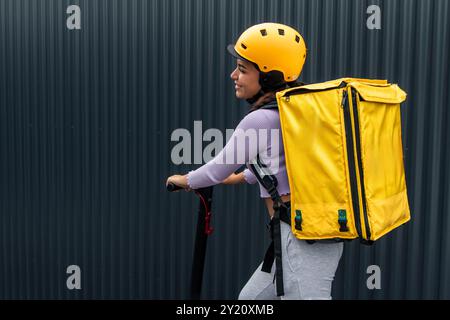 Die lächelnde lateinische Essensliefererin, die einen gelben Rucksack und Helm trägt und einen Elektroroller fährt, wird von hinten an einem schwarzen Zaun gesehen Stockfoto