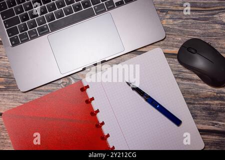 Ein flaches Design mit rotem Notizbuch, Füllfederhalter, Laptop und Maus auf einem rustikalen Holzschreibtisch, das moderne Grundausstattung für den Arbeitsplatz darstellt Stockfoto