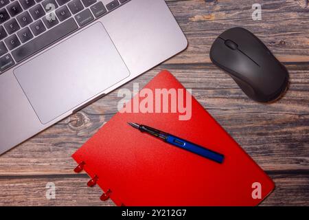 Ein flaches Design mit rotem Notizbuch, Füllfederhalter, Laptop und Maus auf einem rustikalen Holzschreibtisch, das moderne Grundausstattung für den Arbeitsplatz darstellt Stockfoto