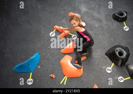 Das kleine Mädchen trainierte klettert in der Kletterhalle an der Wand. Weibliches Kind in Sportbekleidung, das ohne Hilfe im Kinderboulderzentrum arbeitet. Das tapfere und starke Mädchen klettert auf die falsche Wand. Stockfoto