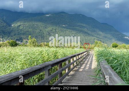 Fußbrücke aus Holz, die zu einer Herzskulptur mit üppigen grünen Bergen und dramatischen Wolken im Hintergrund führt, Liebeskonzept Stockfoto