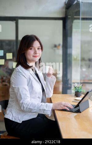 Eine schöne, positive kaukasische Geschäftsfrau arbeitet in einem Café an ihrem digitalen Tablet und lächelt mit einer Hand auf dem Tablet-Schlüssel in die Kamera Stockfoto