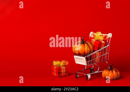 Supermarkt-Trolley mit roten Geschenken mit goldenen Bögen, Einkaufsmöglichkeiten und orangefarbenen Kürbissen. Halloween Shopping und Geschenkkonzept. Roter Hintergrund des Kopierbereichs Stockfoto