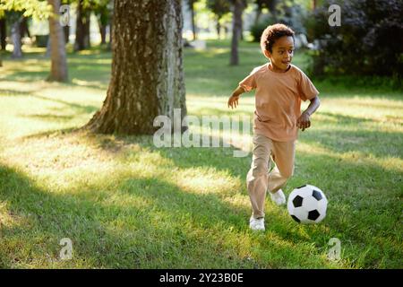 Afroamerikanischer Junge läuft und tritt Ball, während er im Sommerpark abhängt Stockfoto